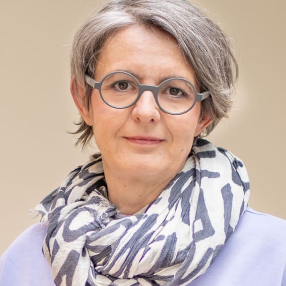 Porträt einer Frau mit kurzen grauen Haaren, runden Brillengläsern und gemustertem Schal